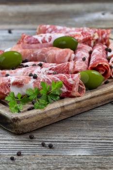 Close up vertical image of various meats on serving board with ham, pork, beef, parsley, and olives on rustic wood. Focus on front top part of serving board and first row of meat. 
