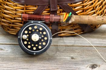 Close up of an antique fly fishing reel, rod, and artificial flies in front of creel with rustic wood underneath. Layout in horizontal format.