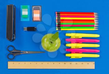 Office or back to school supplies consisting of a green apple, highlight markers, stapler, thumb drive, ruler, scissors, tab markers and colorful pencils on blue background.  