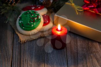 Close up of burning red candle with cookies, present, and evergreen branches on rustic wood. Selective focus on front upper part of candle and flame. 