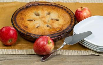 Freshly baked organic apple pie with whole apples, server and plates on brown table cloth.