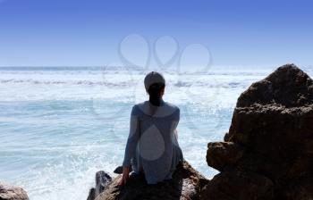 Back view of a woman, sitting on rock, looking outward towards the Pacific Ocean. 