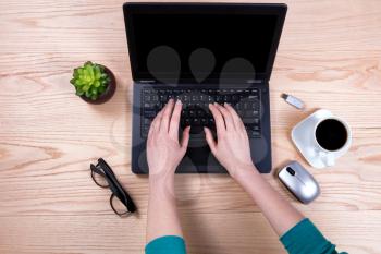 Overhead view of hands typing on laptop keyboard with wireless mouse, baby plant, coffee, thumb drive, and reading glasses on desktop. 