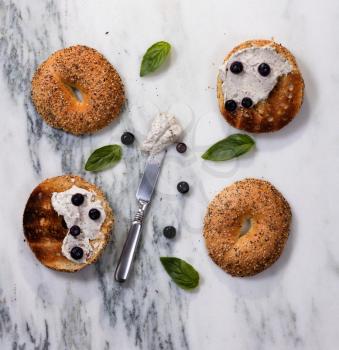 Overhead view of toasted bagels with cream cheese, blueberries, basil, and used knife on white marble stone. 