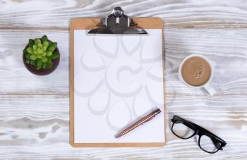Clipboard and pen with reading glasses, plant and creamy coffee on rustic white desktop