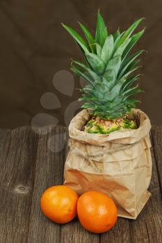 Ripe pineapple in paper bag with oranges on wooden table. Closeup.