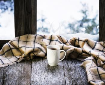 White cup of coffee or tea and woolen plaid located on stylized wooden window sill. Winter concept of comfort and relaxation.