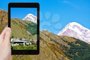 travel concept - tourist takes picture of village Stepantsminda, Gergeti Trinity Church and Mount Kazbek in Caucasus Mountains in Georgia on smartphone,