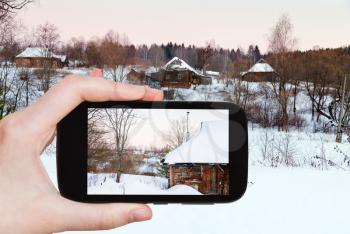 travel concept - tourist takes picture of rural landscape with snowy wooden houses in country at pink winter sunset on smartphone, Russia