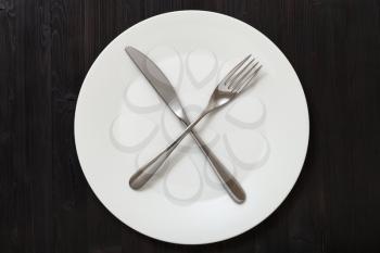 food concept - top view of white plate with crossing knife, spoon on dark brown table