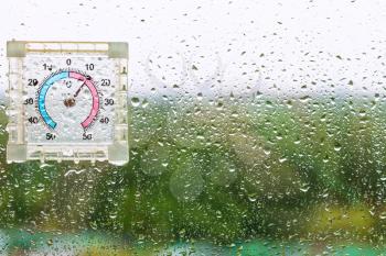 rain drops and wet thermometer on window glass and blurred city park on background on cool rainy autumn day