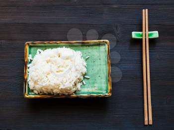 top view of portion of boiled rice on green plate and wooden chopsticks on rest on dark brown table