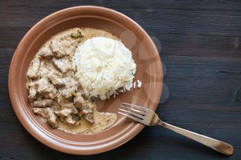 Russian cuisine dish - prepared for eating a portion of Beef Stroganoff (Beef Stroganov, Befstroganov) pieces of stewed meat in sour cream with boiled rice on brown plate on dark table