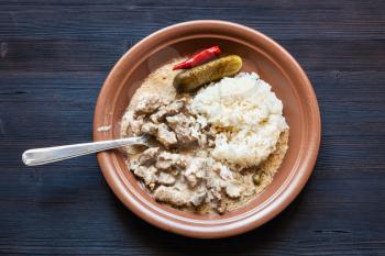 Russian cuisine dish - eating of Beef Stroganoff (Beef Stroganov, Befstroganov) pieces of stewed meat in sour cream with boiled rice on brown plate on dark wooden board