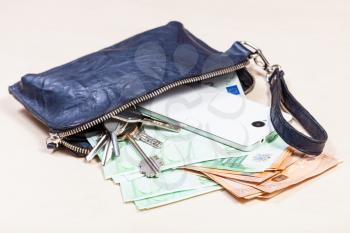 open blue leather wristlet pouch bag with bunch of keys, smartphone and various euro banknotes on pale brown table