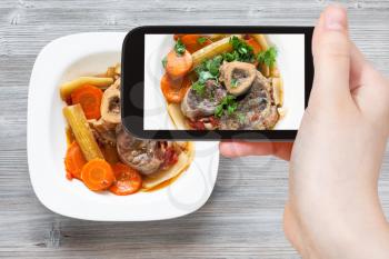 travel concept - visitor photographs of italian dish Ossobuco in white plate on smartphone