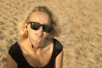 Beautiful young blond girl with glasses and headphones on the background of a sandy beach, edited with sepia