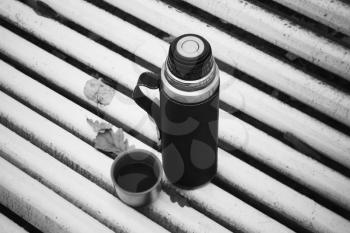 Thermos with cup on wooden bench in autumn park. Black and white photo