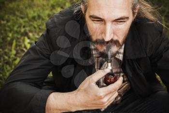 Young serious bearded man smoking pipe in summer park, close up portrait