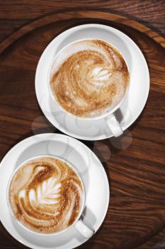 Two cups of cappuccino coffee stand on dark wooden table, close-up vertical photo top view