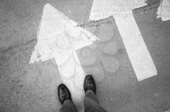 Male feet in new black shining leather shoes stand on asphalt pavement with white arrows pedestrian crossing road marking, first person view