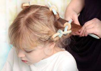 Little blond Russian girl with curlers on hair and hairdressers hands