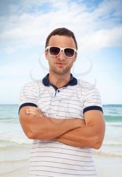 Outdoor portrait of young sporty smiling Caucasian man standing with white sunglasses on summer sea coast