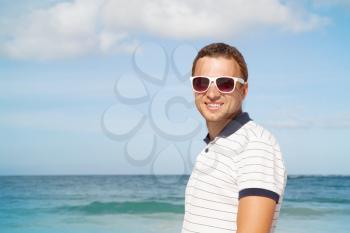Outdoor portrait of young smiling Caucasian man standing with white sunglasses on summer sea coast