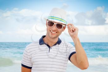 Outdoor portrait of young smiling Caucasian man in white hat and sunglasses standing on the coast