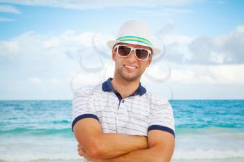 Outdoor portrait of young smiling Caucasian man in white hat and sunglasses standing on the sea coast