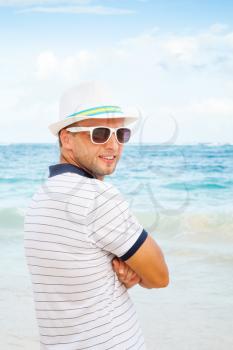 Outdoor portrait of young smiling Caucasian man in white hat and sunglasses standing on the summer ocean coast