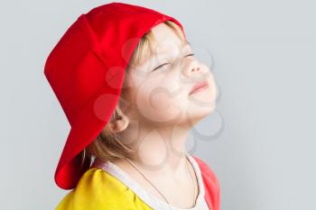 Studio portrait of happy baby girl with closed eyes in red baseball cap over gray wall background
