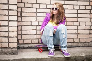 Blond teenage girl in a sunglasses with skateboard sits near urban brick wall, photo with retro tonal correction, instagram old style filter