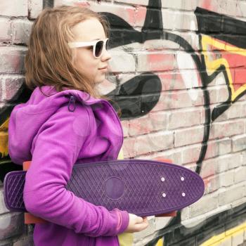 Blond teenage girl holds skateboard, urban brick wall with graffiti on a background
