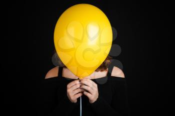 Girl hiding her face under yellow balloon, studio shot over black background