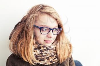 Close up portrait of beautiful blond Caucasian teenage girl in glasses and warm scarf over white wall background
