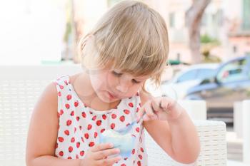Cute blond Caucasian baby girl eats frozen yogurt with ice cream and fruits, close up outdoor portrait with natural high key light