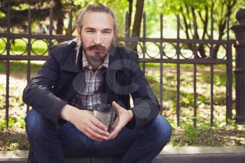 Bearded Asian man sitting on the sidewalk in park with paper cup of coffee