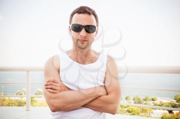 Young sporty Caucasian man in white shirt and black sunglasses stands with crossing hands. Outdoor summer portrait with sea on a background