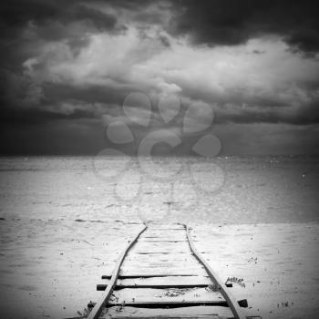Old railway goes over sandy beach to sea water under dark dramatic cloudy sky, useful for boat launching. Square black and white photo with selective focus