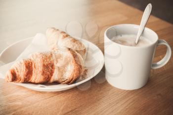 Cappuccino with croissant. White cup of coffee with milk foam stands on wooden table, closeup photo with soft selective focus. Vintage tonal correction filter, old style photo effect
