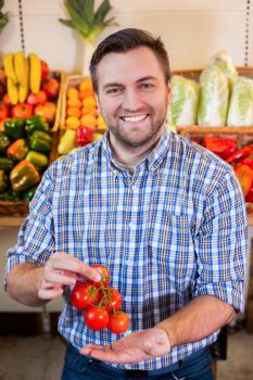Positive man showing ripe tomato branch in market. Grocery on the background.