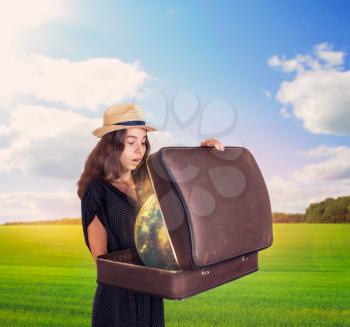 Young woman holding a half-open suitcase with magic globe. Green meadow and the sky with clouds at the background. Travel concept.