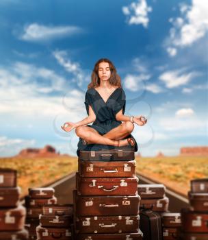 Young girl sits in a pose of yoga on a mountain of suitcases. Blur road and rocky mountains on the background.
