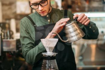 Waiter in black apron and eyeglasses pours hot boiled water in a coffee pot standing on bar counter.