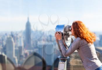 Red-haired young woman looking at the panorama of the city in an observation coin operated binoculars.