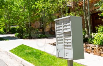 Group of grey metal mailboxes are on the sidewalk with green grass.