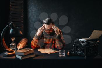 Thoughtful bearded author in glasses reading his work. Retro typewriter, feather, crystal decanter, books and vintage lamp on the desk