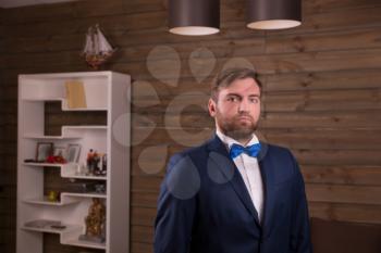 Portrait of serious groom in suit and bow tie, wooden room interior on background