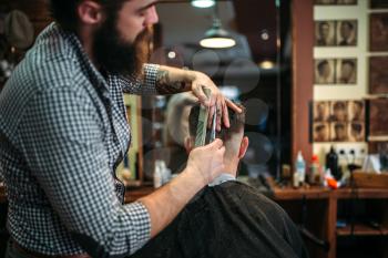 Bearded coiffeur cutting hairstyle by scissors. Client man in black salon cape sitting against a mirror at the barbershop